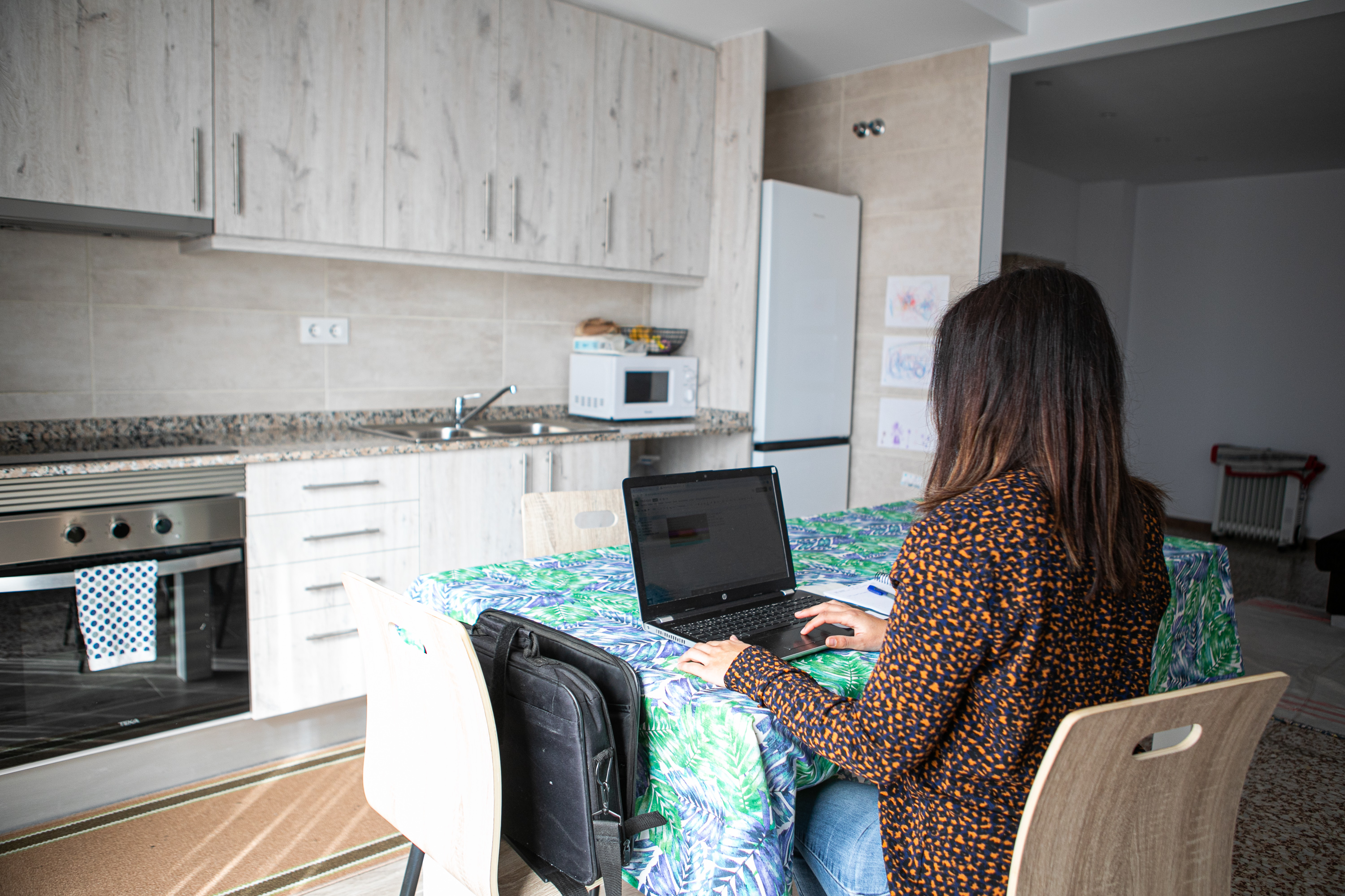 Woman from the back sitting in the kitchen in front of a notebook (2021), (c)Bloc Cooperatiu